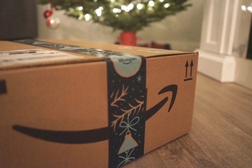 A cardboard box with Amazon branding sits on a wooden floor, with festive blue decorative tape featuring holiday motifs. In the background, a blurred image of a Christmas tree illuminated with lights can be seen, along with a few red and silver ornaments.