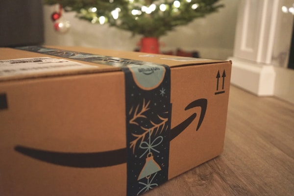 A cardboard box with Amazon branding sits on a wooden floor, with festive blue decorative tape featuring holiday motifs. In the background, a blurred image of a Christmas tree illuminated with lights can be seen, along with a few red and silver ornaments.