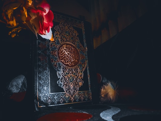 A richly decorated book with intricate designs and Arabic script embossed on the cover. The scene is dimly lit, creating a dramatic contrast with the vibrant artificial flowers in shades of red and yellow in the top left corner.