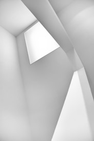 A minimalist architectural composition featuring sharp geometric lines and angles. The play of light and shadows creates a series of intersecting planes and a sense of depth. The use of white and grey tones adds to the abstraction and simplicity.