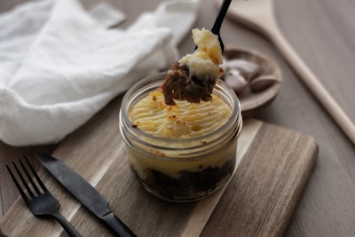 A jar filled with layers of mashed potato and savory meat is placed on a wooden board. A black fork is lifting a portion of the food from the jar. Nearby, there is a folded white napkin and black cutlery, creating a rustic and cozy dining setup.
