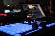 A close-up view of a music production or DJ setup in a dimly lit environment. Illuminated buttons and knobs on a controller are prominently featured, with a laptop displaying audio software in the background. The scene gives a technological and creative vibe.