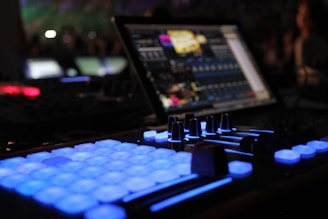 A close-up view of a music production or DJ setup in a dimly lit environment. Illuminated buttons and knobs on a controller are prominently featured, with a laptop displaying audio software in the background. The scene gives a technological and creative vibe.