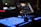 A close-up view of a music production or DJ setup in a dimly lit environment. Illuminated buttons and knobs on a controller are prominently featured, with a laptop displaying audio software in the background. The scene gives a technological and creative vibe.