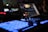 A close-up view of a music production or DJ setup in a dimly lit environment. Illuminated buttons and knobs on a controller are prominently featured, with a laptop displaying audio software in the background. The scene gives a technological and creative vibe.