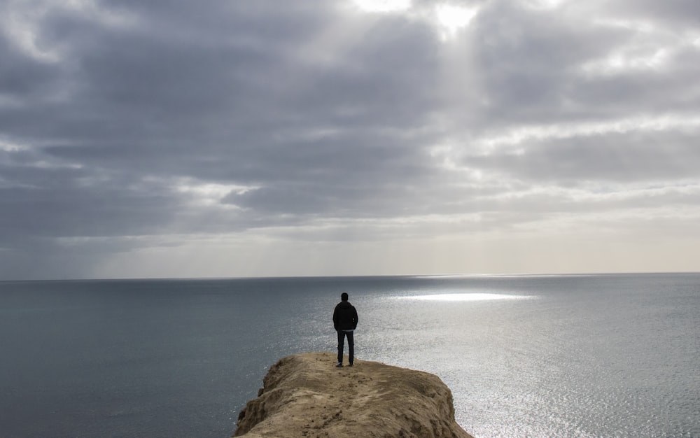 person standing on brown rock formation near body of water under cloudy sky during daytime
