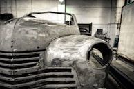 A vintage car is undergoing restoration in an industrial garage setting. The front of the car is stripped to bare metal, showing signs of sanding and preparation work. The environment is dimly lit with scattered tools and equipment visible in the background.