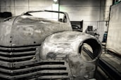 A vintage car is undergoing restoration in an industrial garage setting. The front of the car is stripped to bare metal, showing signs of sanding and preparation work. The environment is dimly lit with scattered tools and equipment visible in the background.
