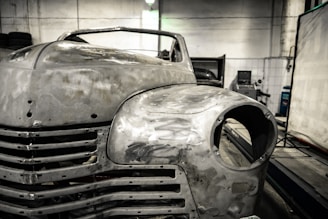 A vintage car is undergoing restoration in an industrial garage setting. The front of the car is stripped to bare metal, showing signs of sanding and preparation work. The environment is dimly lit with scattered tools and equipment visible in the background.
