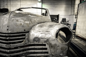 A vintage car is undergoing restoration in an industrial garage setting. The front of the car is stripped to bare metal, showing signs of sanding and preparation work. The environment is dimly lit with scattered tools and equipment visible in the background.