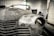 A vintage car is undergoing restoration in an industrial garage setting. The front of the car is stripped to bare metal, showing signs of sanding and preparation work. The environment is dimly lit with scattered tools and equipment visible in the background.