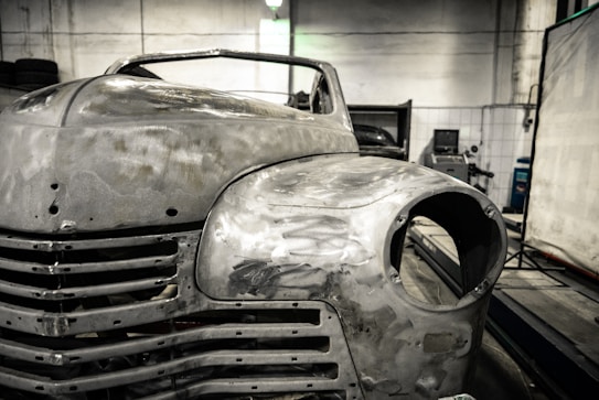 A vintage car is undergoing restoration in an industrial garage setting. The front of the car is stripped to bare metal, showing signs of sanding and preparation work. The environment is dimly lit with scattered tools and equipment visible in the background.