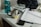 A cluttered desk features a silver metal mug with a teabag attached. Papers, notebooks, and a smartphone are scattered on the surface. In the background, a large blue water bottle and other miscellaneous items are visible.