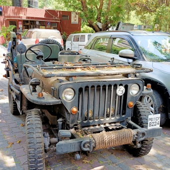 An old, rugged military-style jeep is parked outdoors on a paved area. The vehicle has gathered leaves on its hood, indicating it has been stationary for some time. It features prominent round headlights, a sturdy grill, and a rope coiled at the front bumper. There are other vehicles parked nearby and trees with dense foliage providing shade.