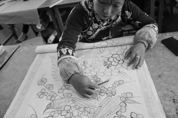 A person engaged in intricate crafting, drawing a detailed floral design on a large piece of fabric. The artwork features flowers and a bird, with precise lines and patterns. The workspace includes a table with tools and other materials in the background.