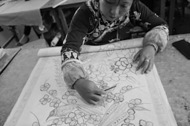A person engaged in intricate crafting, drawing a detailed floral design on a large piece of fabric. The artwork features flowers and a bird, with precise lines and patterns. The workspace includes a table with tools and other materials in the background.