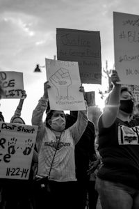 A black and white photograph of a protest where individuals are holding signs with messages advocating for justice, referencing George Floyd and against police brutality. People are wearing masks and appear to be in motion, raising signs that convey messages of activism and solidarity.