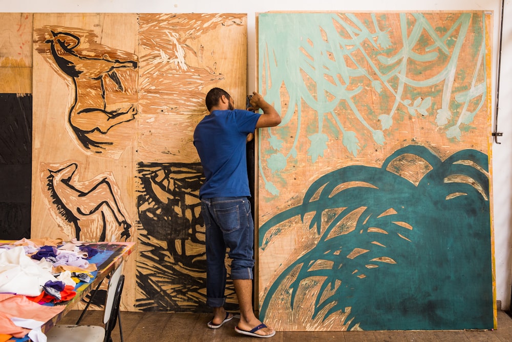 man in blue t-shirt and blue denim jeans standing beside wall with graffiti