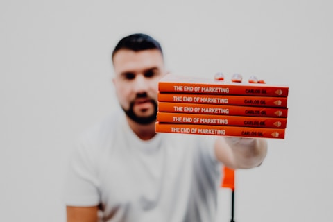 A person in a white t-shirt is holding a stack of five identical red books titled 'The End of Marketing' by Carlos Gil. The background is a very light color, providing minimal distraction from the foreground.