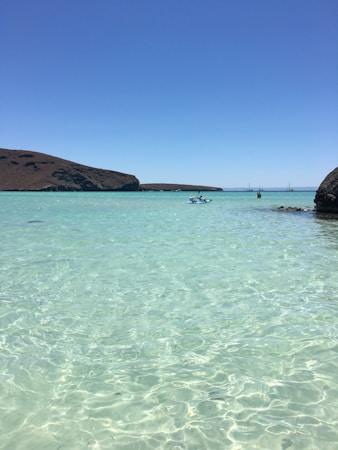 A clear, shallow, turquoise sea bordered by rocky hills under a bright blue sky. Two people are seen kayaking in the water, with several sailboats visible in the distance. The water is calm and crystalline, creating a serene and peaceful atmosphere.