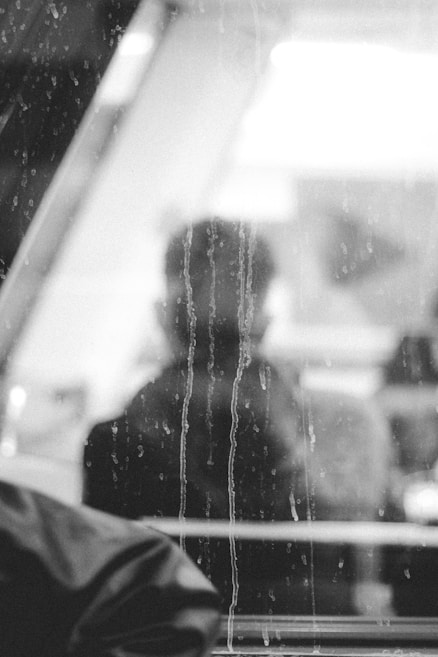 A black and white photograph depicts a blurred silhouette of a person seen through a rain-specked window. The scene creates a feeling of isolation and introspection, enhanced by the focus on the streaks of water on the glass. The background is indistinct, leaving only the blurred contours of the figure.