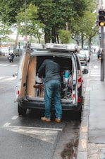 A person is standing at the back of a van parked on the side of the street, organizing or retrieving items. The van's back doors are open, revealing various tools and equipment. The street is lined with trees, and there are several cars and a motorcycle parked along the road. The sidewalk is wet, indicating recent rain.