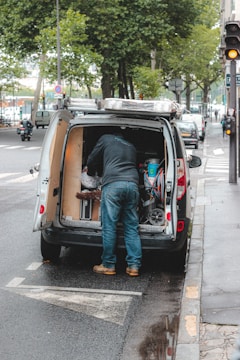 A person is standing at the back of a van parked on the side of the street, organizing or retrieving items. The van's back doors are open, revealing various tools and equipment. The street is lined with trees, and there are several cars and a motorcycle parked along the road. The sidewalk is wet, indicating recent rain.