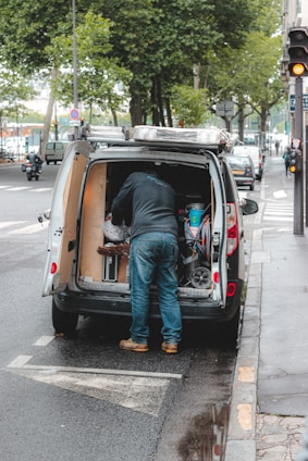A person is standing at the back of a van parked on the side of the street, organizing or retrieving items. The van's back doors are open, revealing various tools and equipment. The street is lined with trees, and there are several cars and a motorcycle parked along the road. The sidewalk is wet, indicating recent rain.