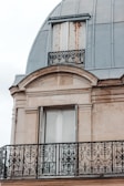 An ornate balcony railing with intricate black wrought iron designs is featured against a beige stone facade. Above, there are closed wooden shutters, slightly worn with rust marks, beneath a dome-shaped metallic roof.