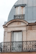 An ornate balcony railing with intricate black wrought iron designs is featured against a beige stone facade. Above, there are closed wooden shutters, slightly worn with rust marks, beneath a dome-shaped metallic roof.