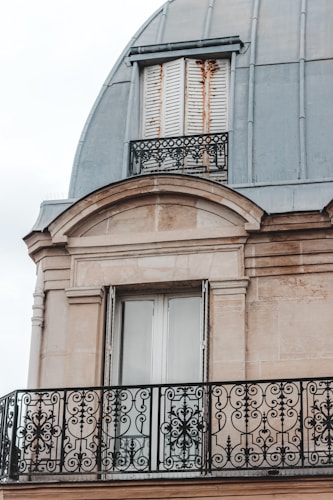 An ornate balcony railing with intricate black wrought iron designs is featured against a beige stone facade. Above, there are closed wooden shutters, slightly worn with rust marks, beneath a dome-shaped metallic roof.