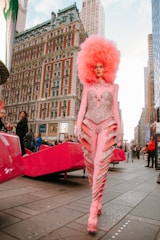 A person walks confidently down a city street wearing an extravagant and glamorous pink outfit adorned with sequins and intricate patterns. The outfit is complemented by a large, voluminous pink afro-style wig. The background features a historic building with detailed architecture, other modern structures, and people observing in the vicinity.