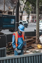 A construction worker wearing a blue uniform and an orange safety vest stands at a work site. The worker is holding a disposable cup and is positioned in front of a blue truck trailer. Various construction materials, including wooden planks and a wire framework, are visible around the site. The background includes urban buildings and a tree, with cars and pedestrians on the street.