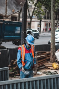 A construction worker wearing a blue uniform and an orange safety vest stands at a work site. The worker is holding a disposable cup and is positioned in front of a blue truck trailer. Various construction materials, including wooden planks and a wire framework, are visible around the site. The background includes urban buildings and a tree, with cars and pedestrians on the street.