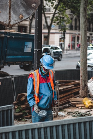 A construction worker wearing a blue uniform and an orange safety vest stands at a work site. The worker is holding a disposable cup and is positioned in front of a blue truck trailer. Various construction materials, including wooden planks and a wire framework, are visible around the site. The background includes urban buildings and a tree, with cars and pedestrians on the street.