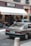 A vintage car is parked on a street in front of a bakery named SECCO, which has a sign for 'Artisan Boulanger Patissier.' The street appears urban, with people visible near the entrance of the bakery, and another car is also partially visible. The façade features balconies with some plants.