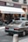 A vintage car is parked on a street in front of a bakery named SECCO, which has a sign for 'Artisan Boulanger Patissier.' The street appears urban, with people visible near the entrance of the bakery, and another car is also partially visible. The façade features balconies with some plants.