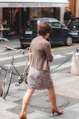 A woman in a floral skirt and brown cardigan is walking across a street. She is wearing brown high heels and holding a beige handbag under one arm. There is a bicycle parked at a street corner secured with a chain. In the background, there are two black cars parked in front of a bakery. Two people can be seen near the bakery entrance, one sitting at a table and the other standing.