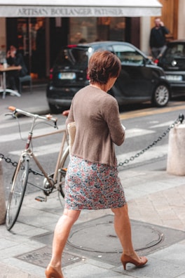A woman in a floral skirt and brown cardigan is walking across a street. She is wearing brown high heels and holding a beige handbag under one arm. There is a bicycle parked at a street corner secured with a chain. In the background, there are two black cars parked in front of a bakery. Two people can be seen near the bakery entrance, one sitting at a table and the other standing.