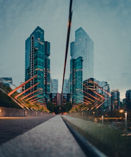 A symmetrical urban scene with tall glass skyscrapers reflecting on a surface, creating an illusion of duplicated buildings. Orange light structures add a vibrant touch, while greenery and a distant tree line the walkway leading towards the cityscape.