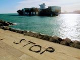 A large container ship marked with 'CMA CGM' navigates through a body of water next to a concrete embankment. The word 'STOP' is spelled out using materials on the embankment, consisting of large stones that form a barrier against the waters.