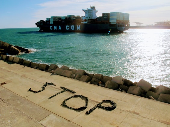 A large container ship marked with 'CMA CGM' navigates through a body of water next to a concrete embankment. The word 'STOP' is spelled out using materials on the embankment, consisting of large stones that form a barrier against the waters.