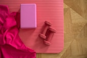 A yoga mat on a wooden floor with a vibrant pink cloth, a pink yoga block, and two red dumbbells placed on it.