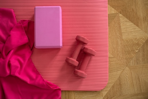 A yoga mat on a wooden floor with a vibrant pink cloth, a pink yoga block, and two red dumbbells placed on it.