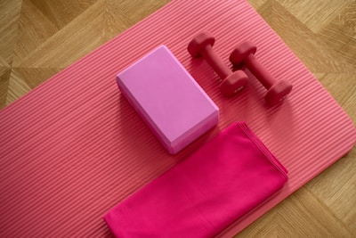 A wooden floor features a pink exercise mat laid out with assorted fitness equipment placed on top. A pink yoga block and a pair of small red dumbbells are neatly arranged. A folded pink towel rests beside them.