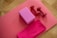 A wooden floor features a pink exercise mat laid out with assorted fitness equipment placed on top. A pink yoga block and a pair of small red dumbbells are neatly arranged. A folded pink towel rests beside them.