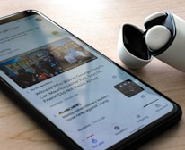 A smartphone on a wooden surface displays a Google News app with an article about George Floyd. Next to the phone, there is a pair of wireless earbuds in a partially opened charging case.