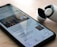 A smartphone on a wooden surface displays a Google News app with an article about George Floyd. Next to the phone, there is a pair of wireless earbuds in a partially opened charging case.