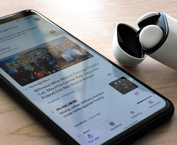 A smartphone on a wooden surface displays a Google News app with an article about George Floyd. Next to the phone, there is a pair of wireless earbuds in a partially opened charging case.