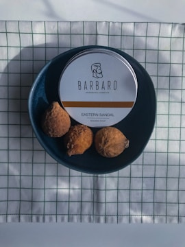 A round tin labeled 'BARBARO Professional Cosmetics' with a logo and the words 'Eastern Sandal Shaving Soap' is placed on a dark blue circular plate. The plate also contains three brown, round objects that appear to be dried fruits or nuts. This setup is on a white cloth with a green grid pattern.